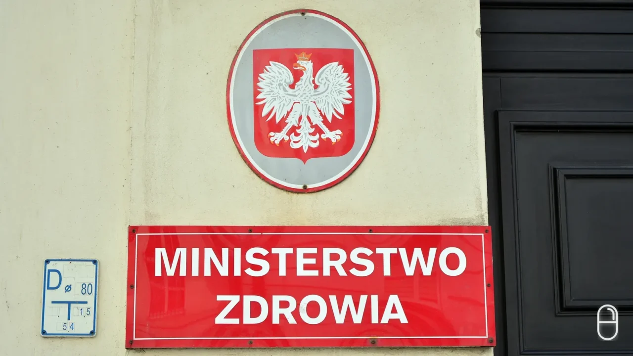 Czerwona tablica z napisem Ministerstwo Zdrowia i godłem Polski wisi na fasadzie budynku obok ciemnych drzwi wejściowych.