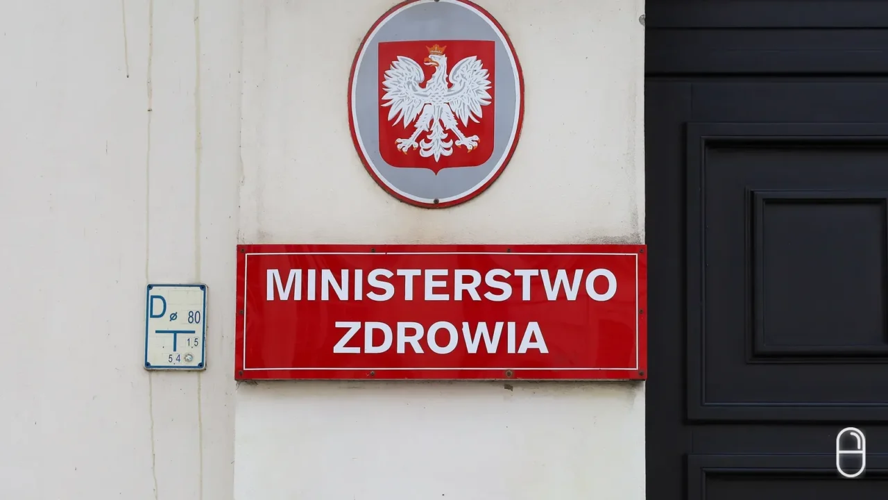 Owalna tablica z godłem Polski i czerwony szyld z napisem Ministerstwo Zdrowia widoczne z bliska przy wejściu do budynku.