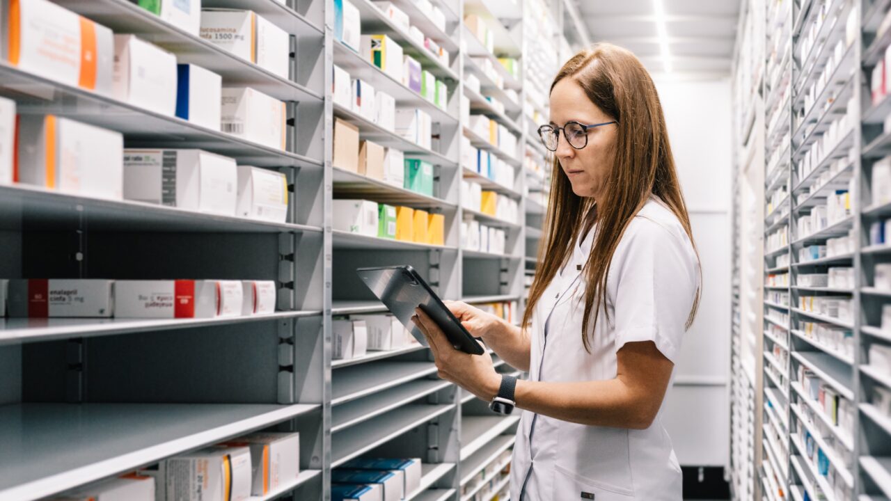 Farmaceuta w białym fartuchu sprawdza stany leków na aptecznych półkach, korzystając z tabletu