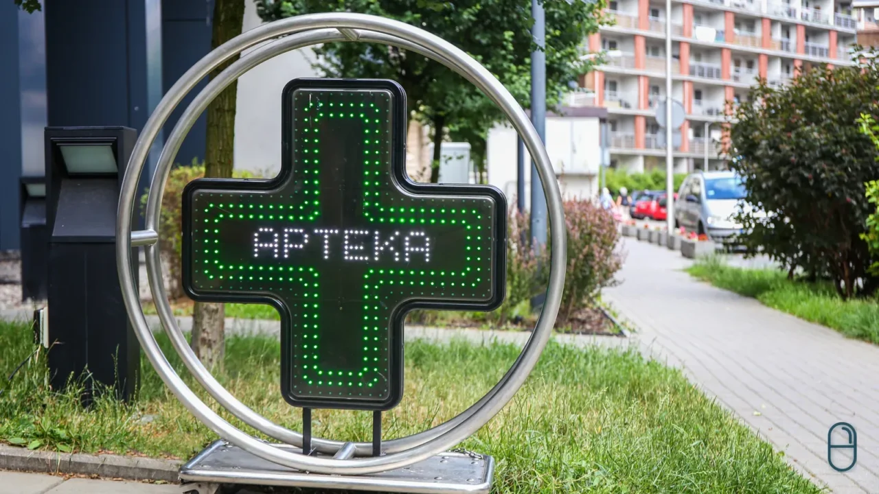 Podświetlony zielony krzyż apteczny z napisem APTEKA wisi w okrągłej metalowej ramie przy chodniku osiedla mieszkaniowego.